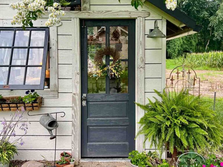 A charming cottage door with a wreath, surrounded by plants, a watering can, and a garden scene in the background. A window with flowers and a hanging lamp are also visible.